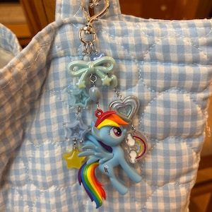 Puede incluir: Un llavero de My Little Pony azul con cola de arcoíris, melena roja y amarilla y alas. El llavero tiene un cierre plateado en forma de corazón y está adornado con abalorios de estrella y lazo. El fondo es un patrón de cuadros azul claro y blanco.