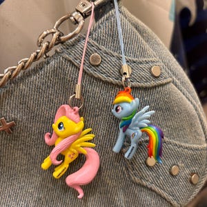 May include: Two My Little Pony character charms attached to a denim bag. One charm is a yellow pony with a pink mane and tail, and the other is a light blue pony with a rainbow mane, tail, and wings. Both figures feature small metal loops.