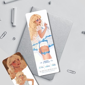 Puede incluir: Un marcap&aacute;ginas con una ilustraci&oacute;n de una mujer rubia sosteniendo un micr&oacute;fono, vestida con un vestido blanco con detalles azules. El marcap&aacute;ginas tiene el texto "The Short N' Sweet Tour" y otros textos. Tambi&eacute;n se ve otro marcap&aacute;ginas con una ilustraci&oacute;n similar.