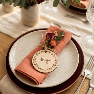 Peut inclure: Mise en place avec une assiette blanche tachetée, une serviette corail et une étiquette de nom en bois avec le mot "Hugo". L'assiette est posée sur une assiette bordeaux, avec une fourchette et un couteau. Un vase de fleurs est en arrière-plan.