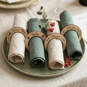 Peut inclure: Mise en place avec des serviettes en lin roulées, crème et vertes, maintenues par des anneaux en bois gravés de noms. Les serviettes sont disposées sur une assiette verte, avec des décorations florales.