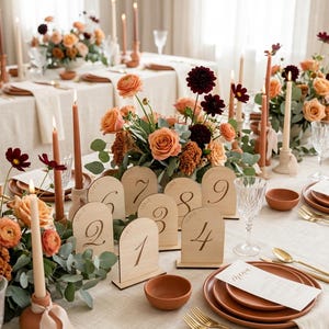 Peut inclure: Un ensemble de numéros de table en bois clair en forme d'arche avec les chiffres un à neuf gravés, disposés sur une table avec des assiettes en terre cuite, des couverts dorés, des bougies effilées et des bouquets de roses orange et dahlias.