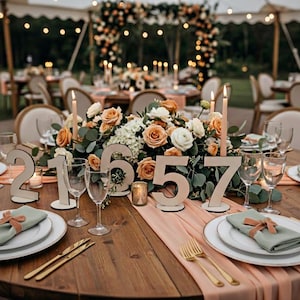 May include: A beautifully decorated wedding reception table with floral centerpieces, candles, and wooden table numbers. The table is set with plates, napkins, and glassware. The color scheme includes peach, white, and green.