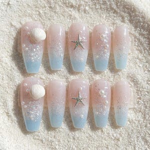 May include: A set of ten artificial nails with a gradient design, transitioning from light pink to light blue with silver glitter. Some nails feature a white seashell, a silver starfish, or small white pearls.