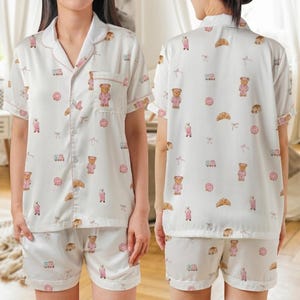 May include: White pajama set with a repeating pattern of pink teddy bears, donuts, croissants, and other small illustrations. The short-sleeved top has a breast pocket with a teddy bear. The shorts have an elastic waistband.