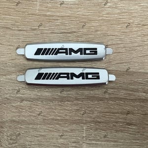 May include: Two silver AMG emblems with black lettering and a distinctive design. The emblems are rectangular with rounded edges and small tabs on each end. The AMG logo is prominently displayed.