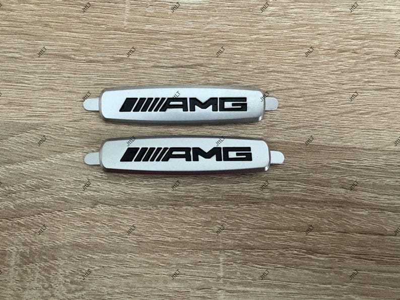 May include: Two silver AMG emblems with black lettering and a distinctive design. The emblems are rectangular with rounded edges and small tabs on each end. The AMG logo is prominently displayed.