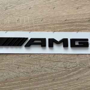 Könnte beinhalten: Schwarzes AMG-Emblem auf weißem Träger. Das Emblem zeigt die Buchstaben "AMG" in einer fetten, serifenlosen Schrift. Das "A" hat eine einfache, blockartige Form, während "M" und "G" stärker stilisiert sind.