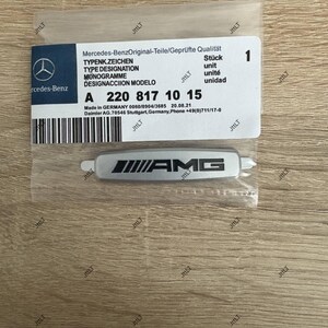 May include: A silver AMG emblem in a clear plastic package, with a white label featuring the Mercedes-Benz logo and product information. The label includes the text "Mercedes-Benz Original-Teile/Gepr&uuml;fte Qualit&auml;t" and the part number A 220 817 10 15.