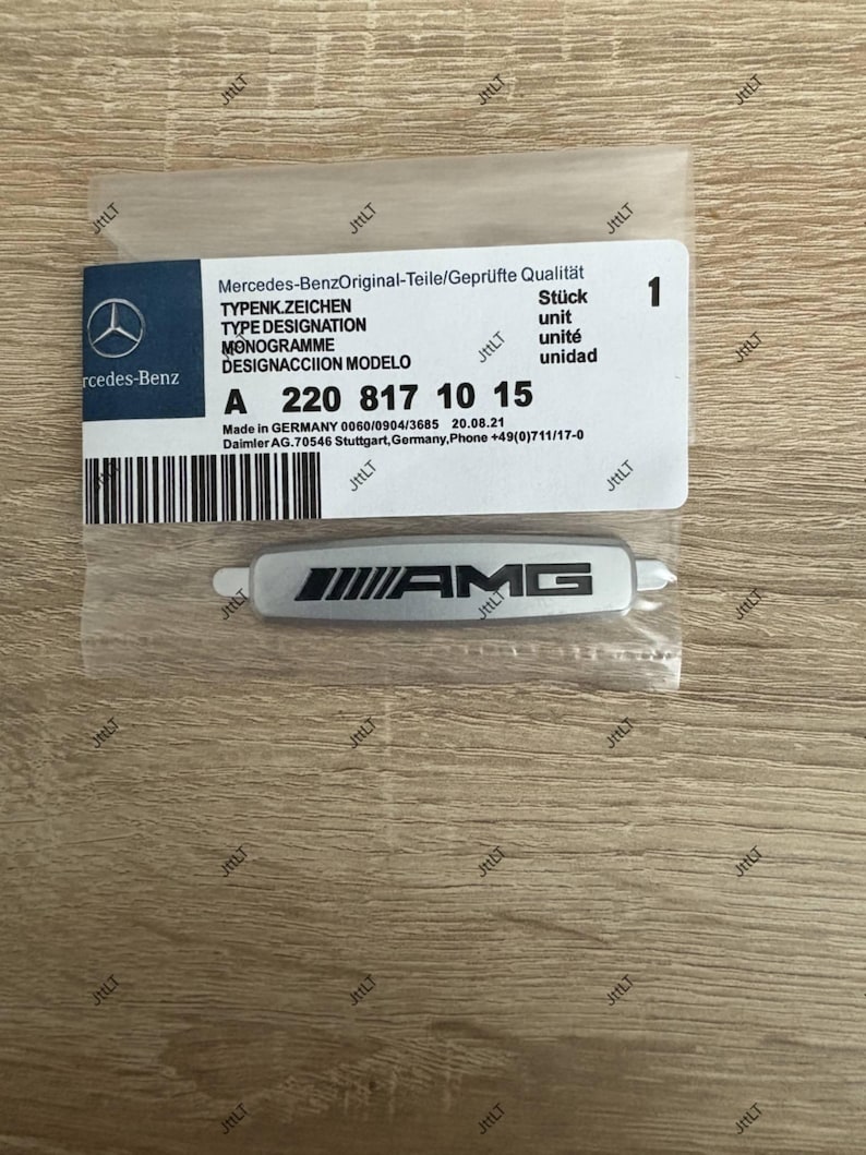 May include: A silver AMG emblem in a clear plastic package, with a white label featuring the Mercedes-Benz logo and product information. The label includes the text "Mercedes-Benz Original-Teile/Gepr&uuml;fte Qualit&auml;t" and the part number A 220 817 10 15.