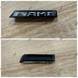 Könnte beinhalten: Zwei schwarze und silberne AMG-Embleme. Das obere Emblem ist rechteckig mit den Buchstaben "AMG" in Silber. Das untere Emblem ist ein schwarzes rechteckiges Stück mit silberner Umrandung.