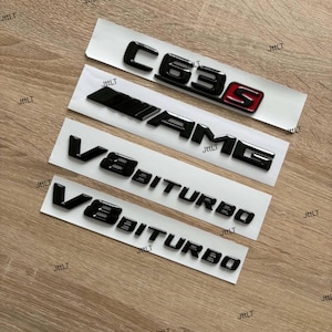 Puede incluir: Un conjunto de cuatro emblemas automotrices negros sobre papel de respaldo blanco. El emblema superior dice C63S con una S roja. El segundo dice AMG. El tercero y cuarto emblemas dicen V8 BITURBO en una fuente negra, elevada y negrita.
