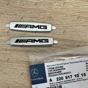 May include: Two silver AMG emblems with black lettering and a Mercedes-Benz parts label. The emblems are rectangular with rounded ends. The label includes the Mercedes-Benz logo and part number A 220 817 10 15.