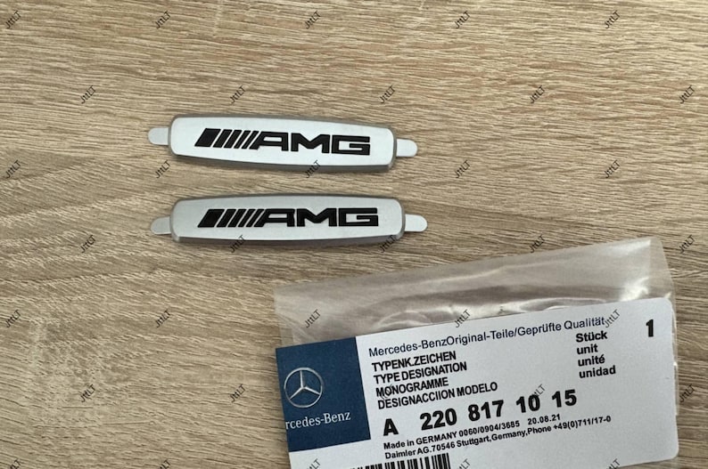 May include: Two silver AMG emblems with black lettering and a Mercedes-Benz parts label. The emblems are rectangular with rounded ends. The label includes the Mercedes-Benz logo and part number A 220 817 10 15.
