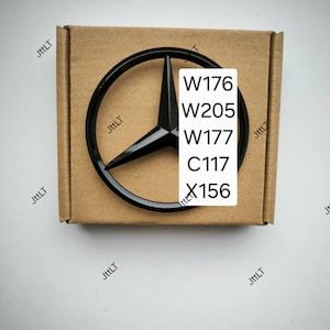 Könnte beinhalten: Ein glänzend schwarzes, kreisförmiges Mercedes-Benz-Emblem mit einem dreizackigen Stern, zentriert auf einer quadratischen braunen Pappschachtel. Ein weißes rechteckiges Etikett ist auf dem Emblem angebracht und zeigt den Text W176, W205, W177, C117 und X156.