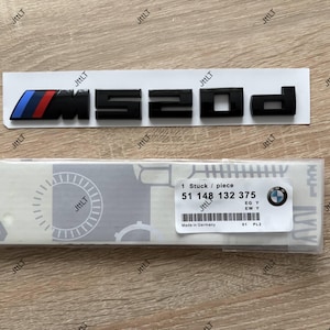 Könnte beinhalten: Schwarzes "M520d"-Emblem mit blauen, roten und schwarzen Streifen. Das Emblem befindet sich auf einem weißen Papierstreifen. Eine Verpackung mit BMW-Logo und dem Text "Made in Germany" ist ebenfalls sichtbar.