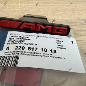 Könnte beinhalten: Ein schwarzes und rotes AMG-Emblem, wahrscheinlich für ein Mercedes-Benz-Fahrzeug, mit dem AMG-Logo. Das Emblem wird von einem Etikett mit Text wie "Mercedes-Benz" und Teilenummern begleitet.