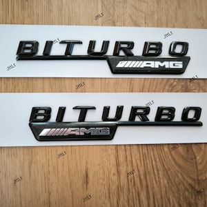 Puede incluir: Dos emblemas BITURBO AMG negros sobre un respaldo blanco. Los emblemas tienen un acabado brillante y están colocados sobre un fondo claro de vetas de madera. El logotipo de AMG es en plata y negro.