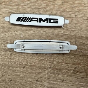 May include: Two silver and white AMG car emblems. The top emblem is white with black AMG lettering. The bottom emblem is silver with two screws. Both emblems have a rectangular shape with rounded edges.