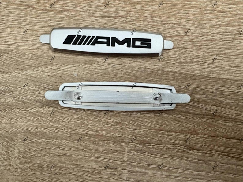 May include: Two silver and white AMG car emblems. The top emblem is white with black AMG lettering. The bottom emblem is silver with two screws. Both emblems have a rectangular shape with rounded edges.