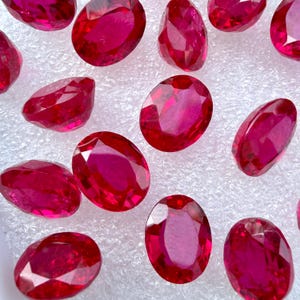 Pode incluir: Uma coleção de pedras preciosas vermelhas vibrantes, com corte oval e superfícies facetadas que refletem a luz. Estas pedras soltas e polidas apresentam um tom carmesim profundo e estão dispostas num padrão disperso.