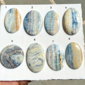 May include: Eight oval-shaped polished gemstones arranged on a white board, numbered one through eight. Each stone features unique banded patterns in shades of blue, white, cream, and orange, with smooth, glossy surfaces and distinct mineral layers.