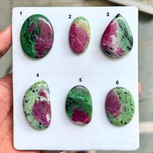 May include: Six polished zoisite gemstones with ruby inclusions arranged on a white display board. Each stone features a unique pattern of vibrant magenta-pink ruby patches set against a mottled green and black zoisite matrix, numbered one through six.