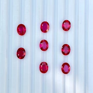 May include: Seven oval-cut, vibrant red gemstones arranged in a grid-like pattern. Each stone features a faceted surface that reflects light, creating a brilliant appearance. The gems are displayed against a white, vertically ribbed background.
