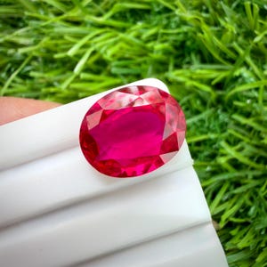 May include: A faceted, oval-shaped ruby gemstone. The gem is a vibrant, deep red color with a high degree of clarity. The ruby is set against a white surface, with a background of green grass.