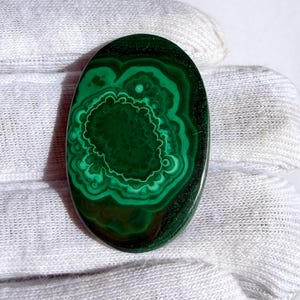 May include: Oval-shaped malachite gemstone with concentric patterns of dark and light green. The polished stone displays a central, textured area surrounded by swirling bands, creating a natural, organic design. The gemstone is set against a white background.
