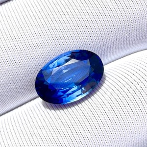 May include: A deep blue, oval-cut gemstone, displayed against a white, textured background. The facets of the gem catch the light, showcasing its colour and clarity. The stone's shape and colour suggest it could be a sapphire.