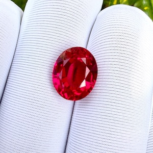 Könnte beinhalten: Ein lebendiger, oval geschliffener, roter Edelstein. Der Edelstein wird vor einem weißen, strukturierten Hintergrund gehalten. Die Facetten des Steins fangen das Licht ein und erzeugen ein brillantes Farb- und Funkeln. Dieser Edelstein ist wahrscheinlich für Schmuck.