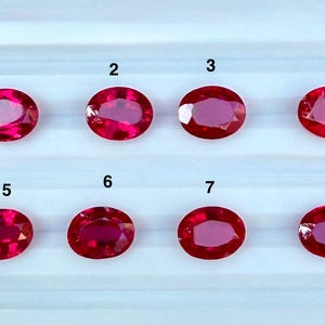 May include: Eight oval-cut, vibrant red gemstones arranged in two rows of four. Each stone is faceted and numbered from one to eight in black text above it. The gems exhibit a consistent deep crimson hue and a polished, reflective surface finish.