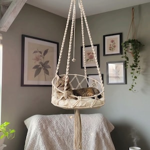 May include: A cream-colored macrame hanging cat bed featuring a circular base with a soft cushion, intricate knotted rope sides, and long twisted suspension cords that meet at a top ring, with a tabby cat resting inside the woven structure.