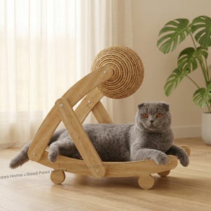 以下が含まれることがあります： 木製の猫用ベッドでくつろぐ灰色の猫。ベッドには編みロープのボールが付いています。ベッドは明るい色の木材でできており、車輪が付いた曲線的なデザインです。猫の毛皮は灰色で、オレンジ色の目をしています。背景には植物があります。