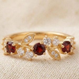 May include: A gold-colored ring featuring a leaf design with red and white gemstones. The ring has a band with a series of small, clear gemstones and larger, round red gemstones. The ring is set against a neutral background.