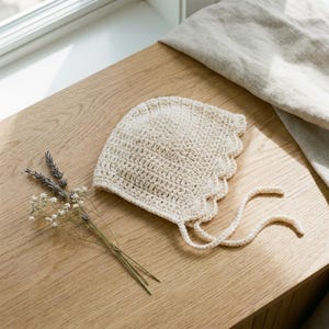 May include: A cream-colored crocheted bonnet featuring a scalloped edge and two long matching ties. The bonnet is crafted with a textured stitch pattern and is accompanied by a small sprig of dried lavender and white baby's breath flowers.