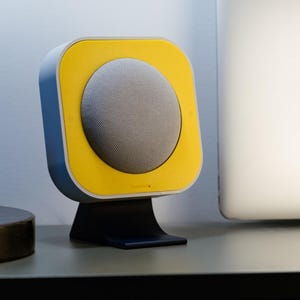 May include: A modern, compact speaker with a yellow and gray design. The speaker has a rounded, mesh-covered front and a square yellow frame. It sits on a black stand, and is likely used for playing music or audio.