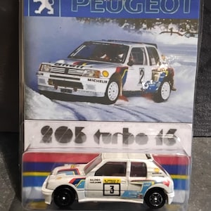 Peut inclure: Modèle réduit d'une Peugeot 205 Turbo 16 blanche, présentée dans un emballage plastique transparent. L'emballage présente le logo Peugeot et une image de la voiture en action. La voiture miniature a des roues noires et des autocollants de course.