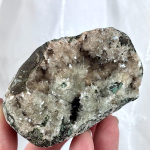 May include: A natural geode with a rough, dark exterior and a cavity filled with sparkling, light-colored crystals. The geode has areas of green and brown hues, and is held in a hand.