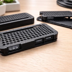 Könnte beinhalten: Schwarze Handyhüllen mit Wabenmuster. Die Hüllen haben Aussparungen für Kamera, Tasten und Ladeanschluss. Sie bestehen aus einem strukturierten Material und liegen auf einer hellen Holzoberfläche.