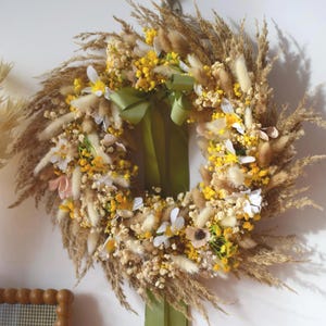 Peut inclure: Une couronne circulaire de fleurs séchées, composée d'herbes, de fleurs jaunes et blanches, et d'un ruban vert. La couronne est suspendue à un mur blanc. Un cadre en bois est visible en bas à gauche.