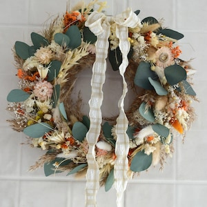 Può includere: Una ghirlanda circolare composta da fiori secchi e verde, tra cui foglie di eucalipto e fiori arancioni. Un nastro color crema con un motivo a quadri è legato in alto, con lunghe code. La ghirlanda è appesa a una parete piastrellata bianca.