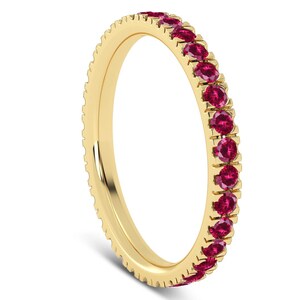 Könnte beinhalten: Ein goldener Bandring mit einer durchgehenden Reihe kleiner, runder, roter Edelsteine. Der Ring hat einen strukturierten Rand und ist vor einem weißen Hintergrund platziert. Der Ring ist ein Schmuckstück.