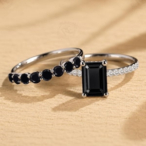 May include: A silver-toned ring set featuring an emerald-cut black center stone flanked by small white accent stones on the band, paired with a matching band adorned with a row of round black stones set in a polished silver-toned metal finish.