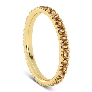 Puede incluir: Un anillo de oro con una banda de pequeñas piedras preciosas redondas de color marrón claro. El anillo tiene un borde texturizado y un acabado dorado pulido. Las piedras están espaciadas uniformemente.