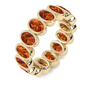May include: A gold ring featuring a series of oval-shaped, orange gemstones. The gemstones are set in a continuous pattern around the ring's band. The ring is set against a white background, highlighting the ring's design.
