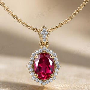 Puede incluir: Un collar con colgante de oro que presenta una piedra preciosa de color rojo intenso con corte ovalado, rodeada por un halo de pequeños diamantes blancos. El colgante cuelga de una fina cadena de oro con diamantes incrustados en el engaste.