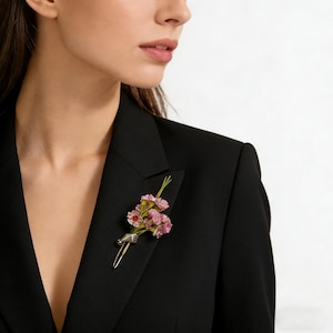 Puede incluir: Broche floral con pequeñas flores rosas y un acento rojo, sujeta a un alfiler de metal plateado. La broche está prendida a una blazer negra, creando un accesorio elegante.