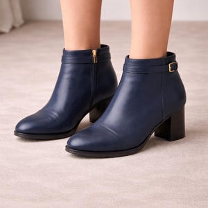 May include: Navy blue ankle boots with a rounded toe and a block heel. The boots feature a side zipper with a gold-tone pull and a decorative strap with a gold-tone buckle. The boots are made of a smooth leather-like material.